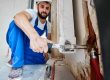 Bearded young man in work overalls using plumber wrench while installing heating radiator in room. Male plumber in safety helmet installing heating system in apartment. Focus on hand with instrument.