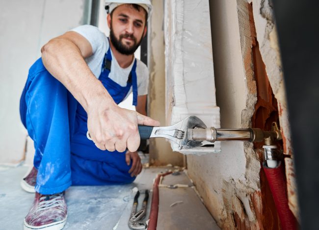 159932996_m Bearded young man in work overalls using plumber wrench while installing heating radiator in room. Male plumber in safety helmet installing heating system in apartment. Focus on hand with instrument.