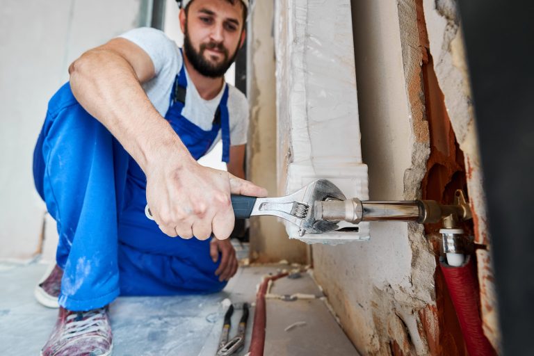 Bearded young man in work overalls using plumber wrench while installing heating radiator in room. Male plumber in safety helmet installing heating system in apartment. Focus on hand with instrument.